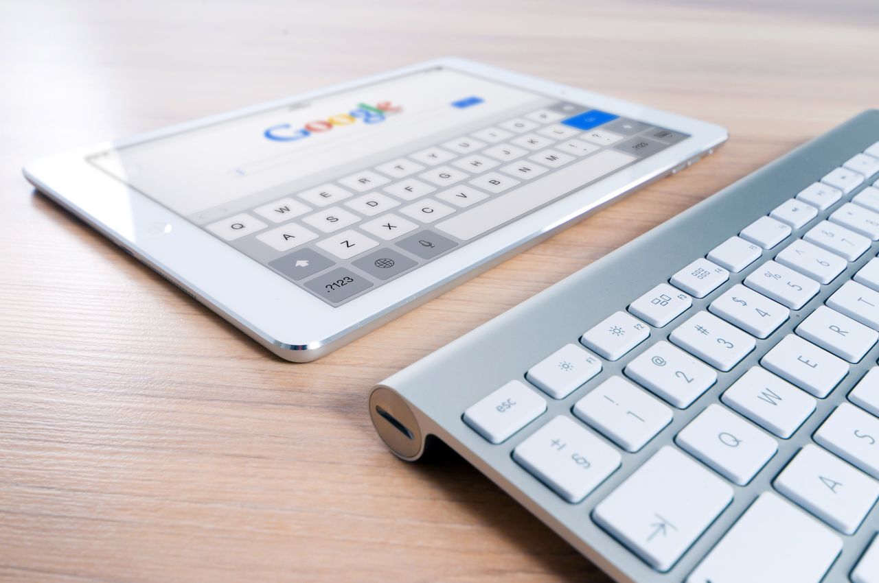 hero-img-02 A modern tablet displaying a search engine logo next to a wireless keyboard on a wooden desk.